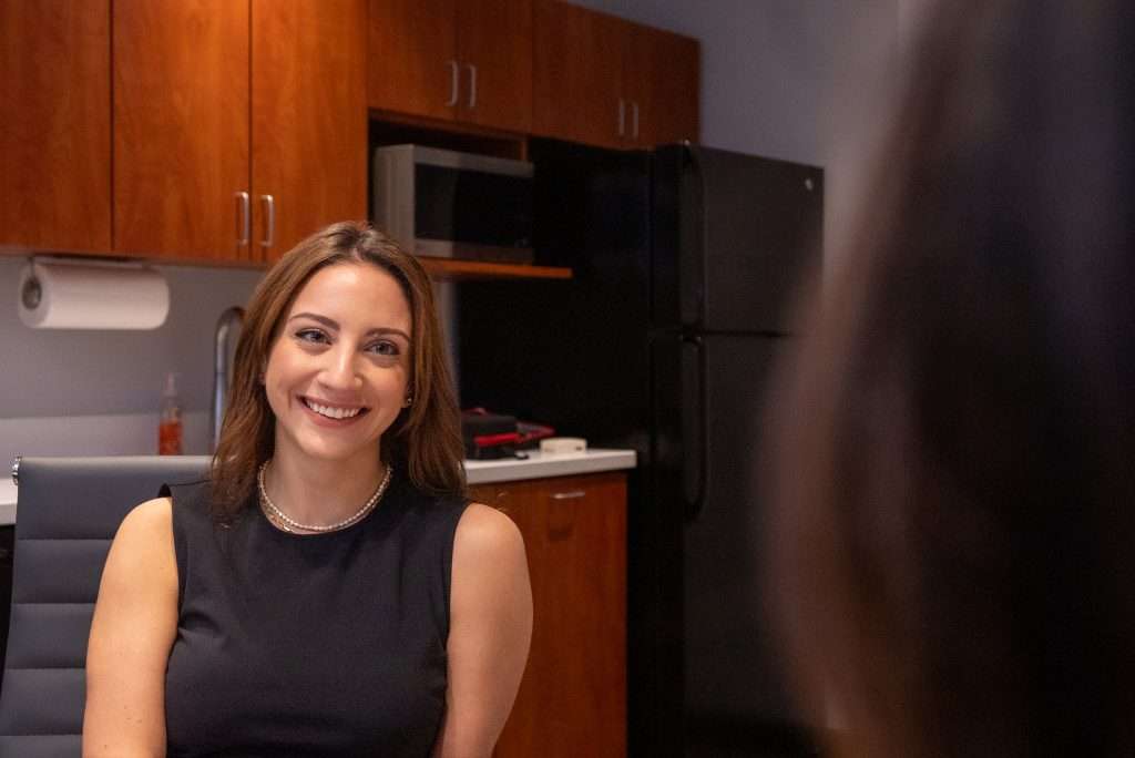 A woman with long brown hair, wearing a sleeveless black dress and a pearl necklace, smiles while sitting in an office kitchen in Washington DC with wooden cabinets, a microwave, and a black refrigerator in the background.
