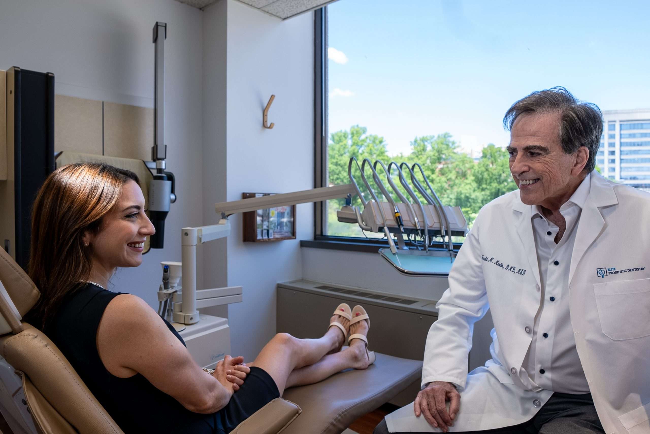 A woman sits in a dental chair smiling and talking with a male dentist in a white coat, inside a bright, modern dental office in Washington DC. The setting suggests a welcoming place for cosmetic dentistry or full mouth reconstruction.